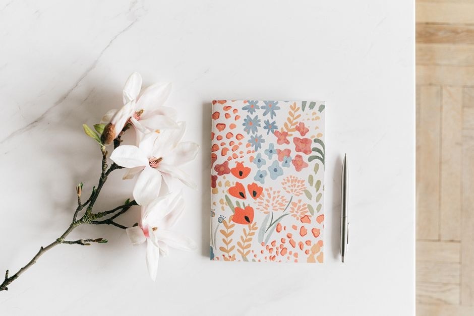 A patterned notebook and pen arranged beside pale flowers on a bright desk.