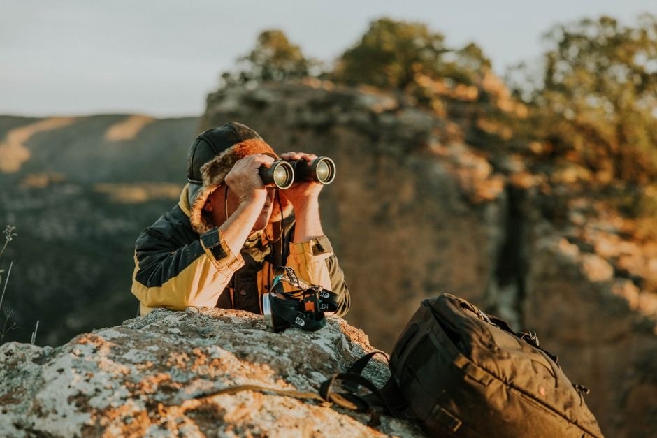 A traveler with binoculars and camera gear pauses on a rocky overlook in warm evening light.